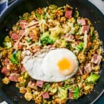 A large dark skillet with a ham breakfast hash topped with shredded cheese and a fried egg with a text overlay on the image
