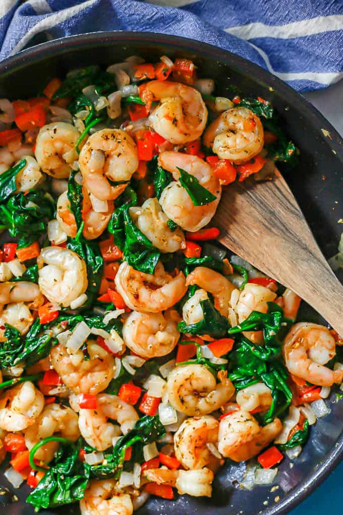 A wooden spatula resting in a large dark skillet with shrimp mixed with sautéed spinach and veggies.