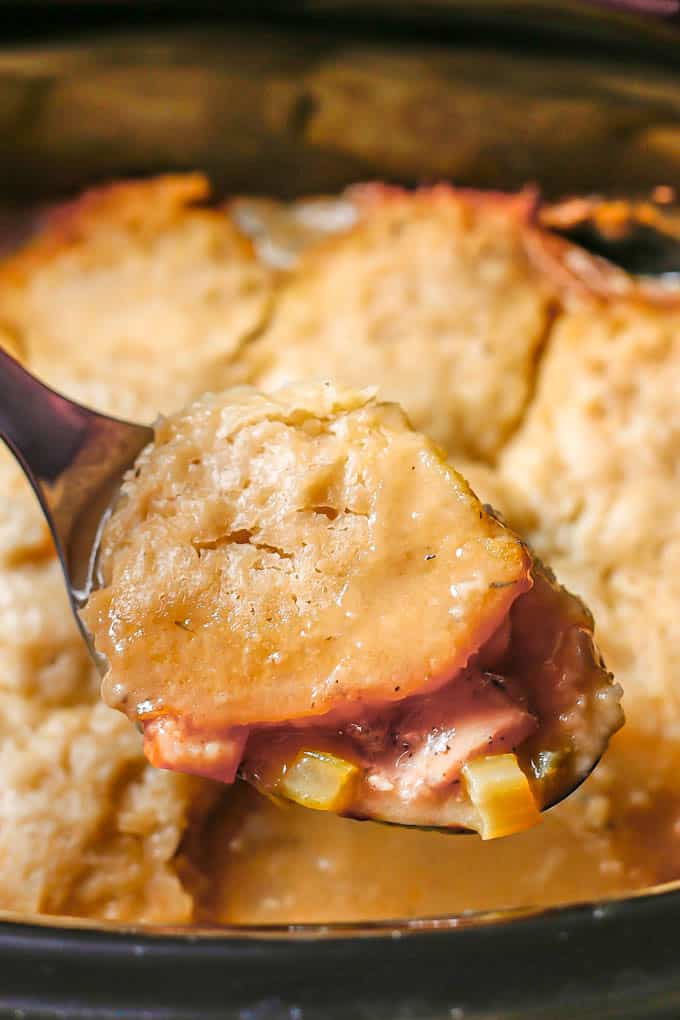 A silver spoon lifting up a scoop of chicken and dumplings from a slow cooker insert.