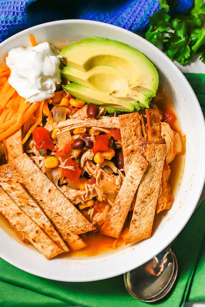 Brothy soup with corn, beans, tomatoes and shredded chicken in a white bowl with homemade tortilla strips on top and garnishes of avocado, Greek yogurt and shredded cheddar