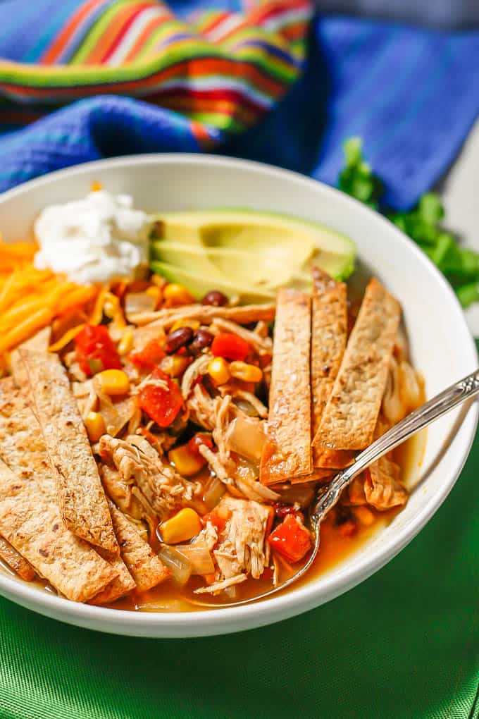 A spoon resting in a white bowl of chicken tortilla soup with avocado, cheese and sour cream for garnishes