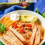 Chicken tortilla soup in a low white bowl with crispy tortilla strips, cheese, sour cream and avocado on top with a text overlay on the image