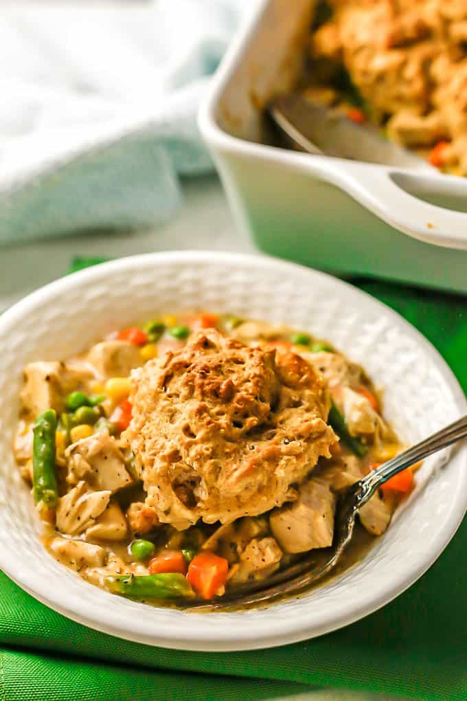 Turkey pot pie with mixed vegetables and a biscuit on top served in a white bowl with the casserole in the background