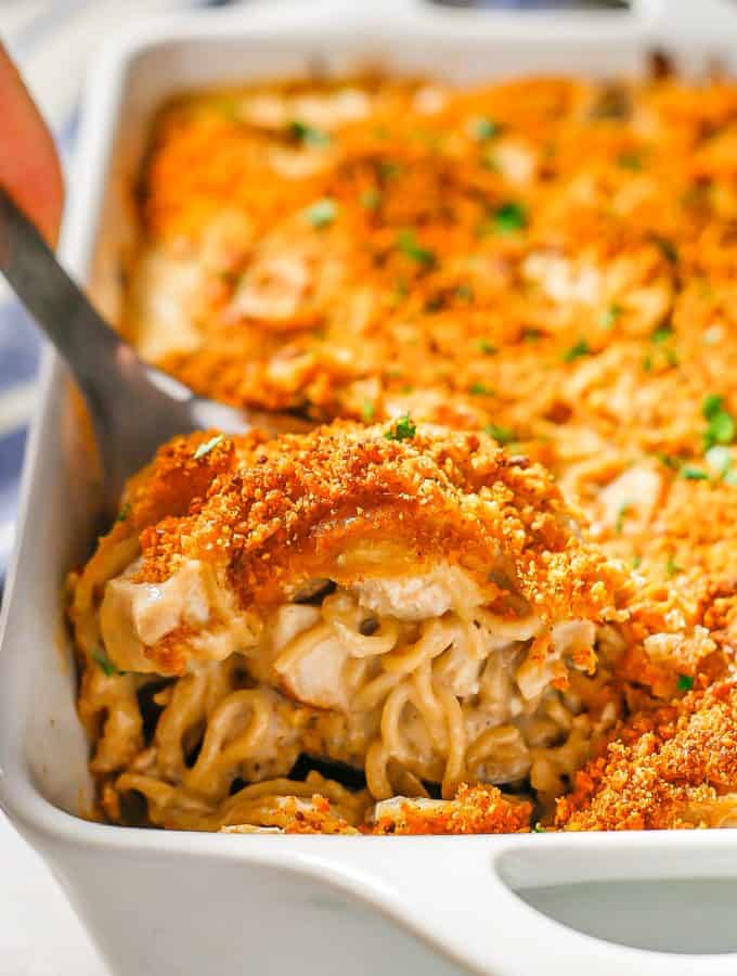 A silver spoon scooping up some creamy turkey tetrazzini with a breadcrumb topping from a white rectangular casserole dish