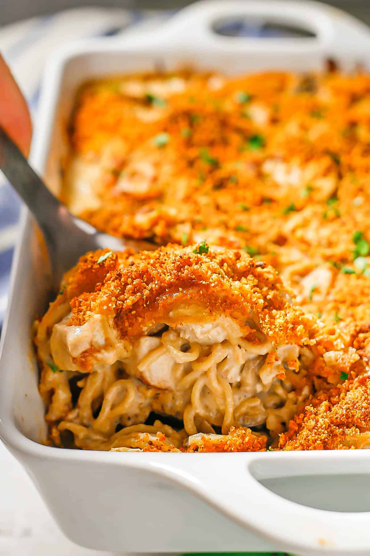 A silver spoon scooping up some creamy turkey tetrazzini with a breadcrumb topping from a white rectangular casserole dish