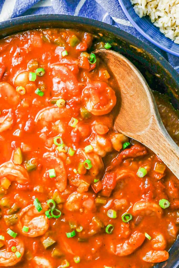 A wooden spoon resting in a skillet with a saucy tomato shrimp mixture topped with sliced green onions.