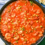 A large dark skillet with shrimp simmered in a tomato sauce and topped with green onions with a text overlay on the photo.