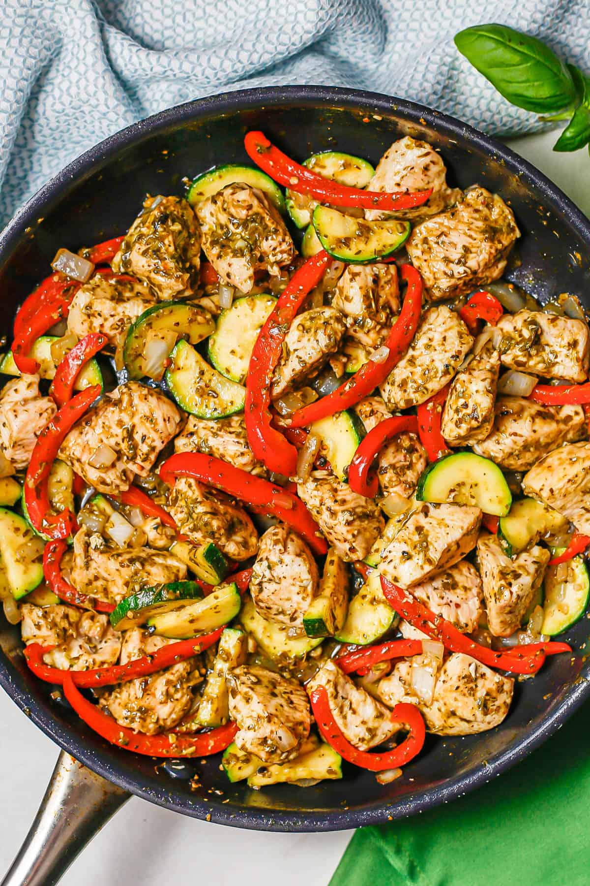 A large skillet with chicken, peppers, zucchini and onion coated in pesto.