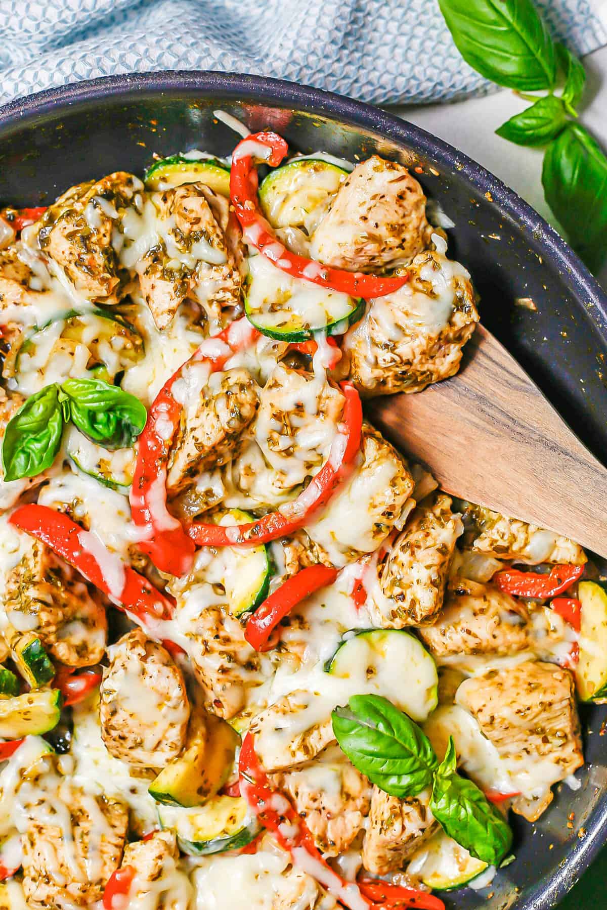 A wooden spatula scooping up a pesto chicken and veggie mixture from a large dark skillet.