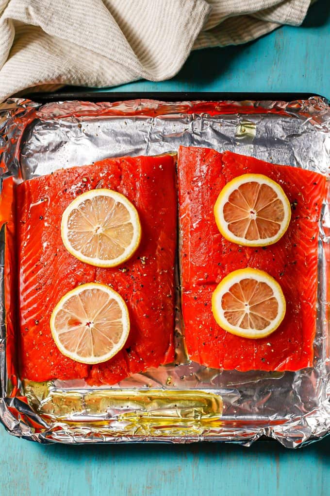 Two salmon filets on a foil lined baking sheet topped with lemon slices before being cooked.