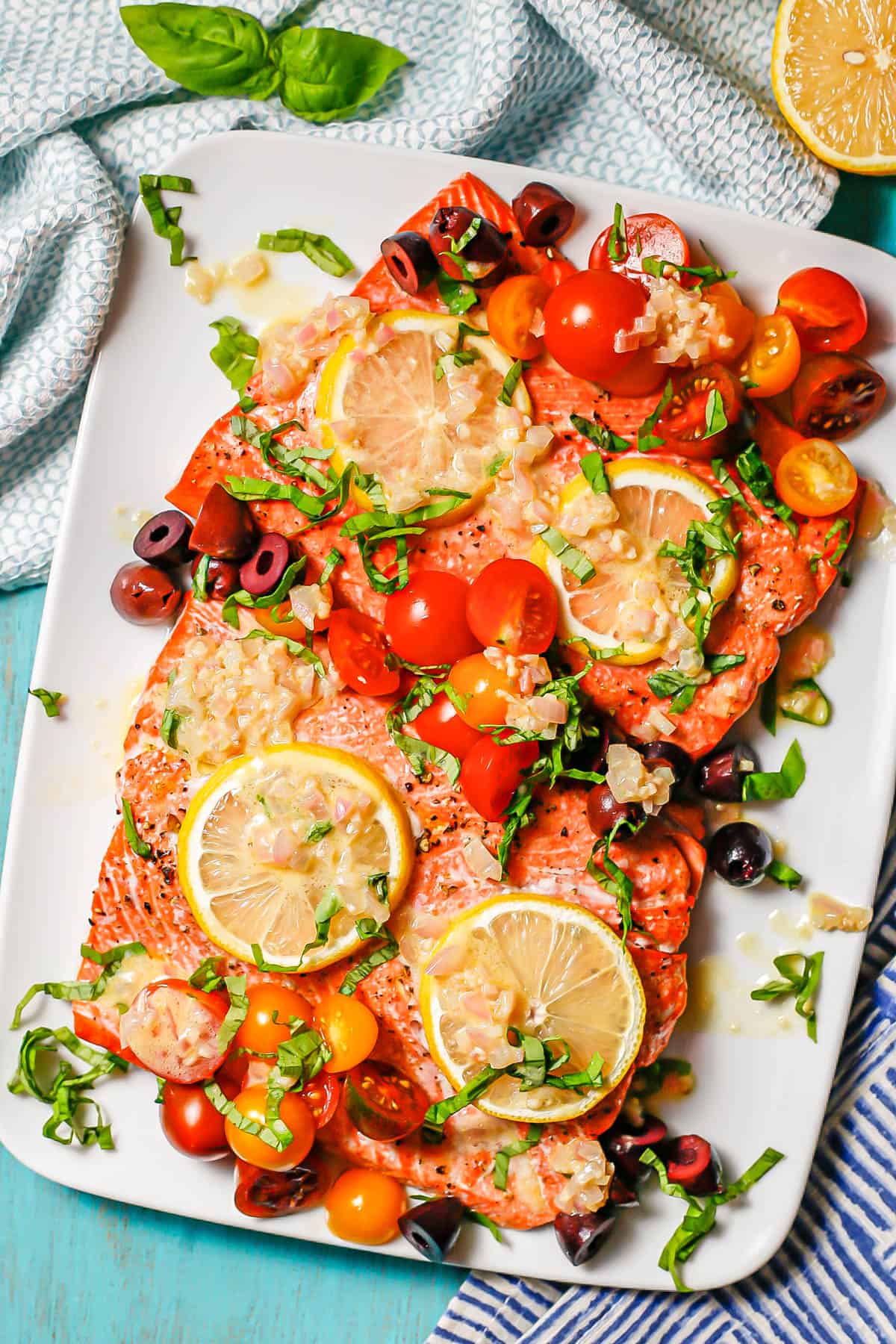 Mediterranean salmon topped with tomatoes, olives, lemon and basil with shallot dressing served on a rectangular white platter with blue napkins nearby.