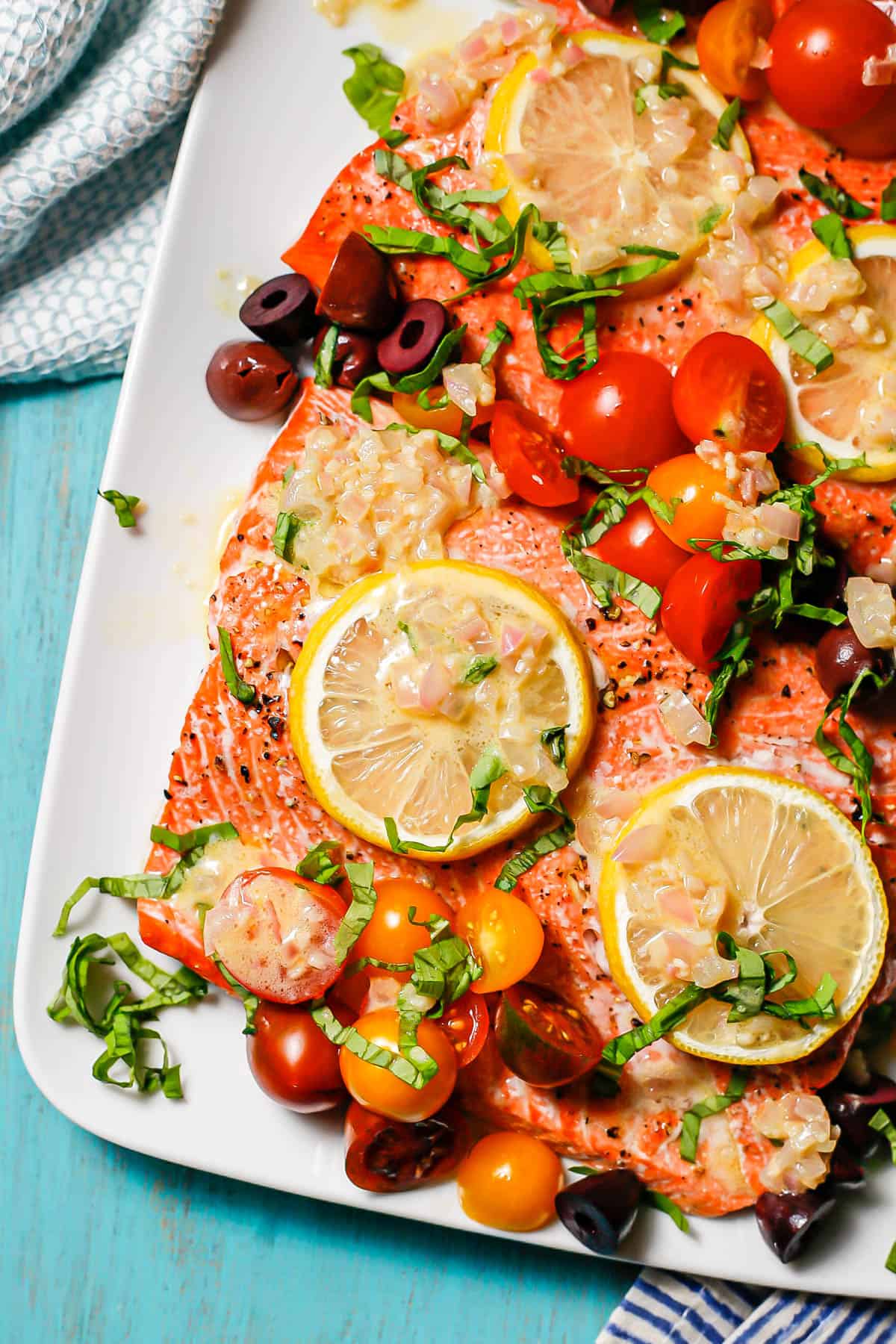 Close up of Mediterranean salmon topped with tomatoes, olives, lemon and basil with shallot dressing served on a rectangular white platter.