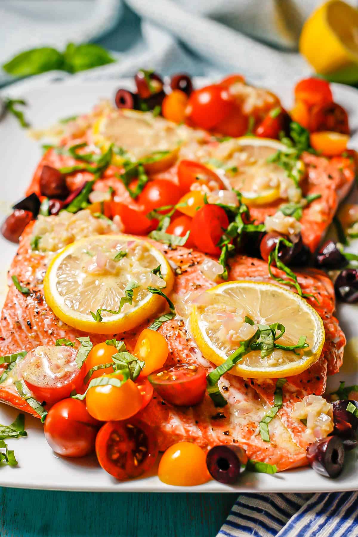 Large salmon filets served on a white platter topped with lemon slices, assorted colored tomatoes, olives, basil and a shallot dressing.