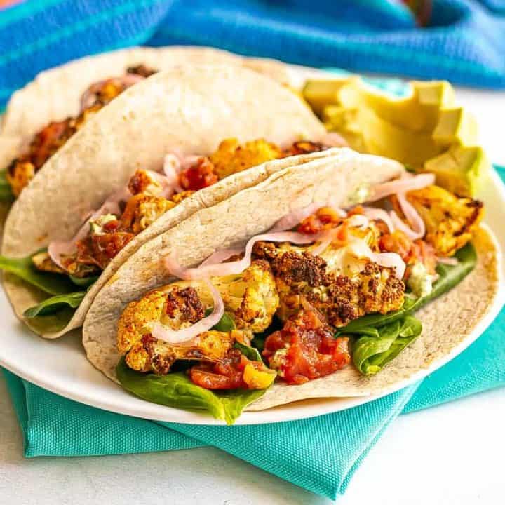 A trio of tacos on a white plate filled with roasted cauliflower florets, spinach, salsa and pickled red onions with avocado slices to the side.