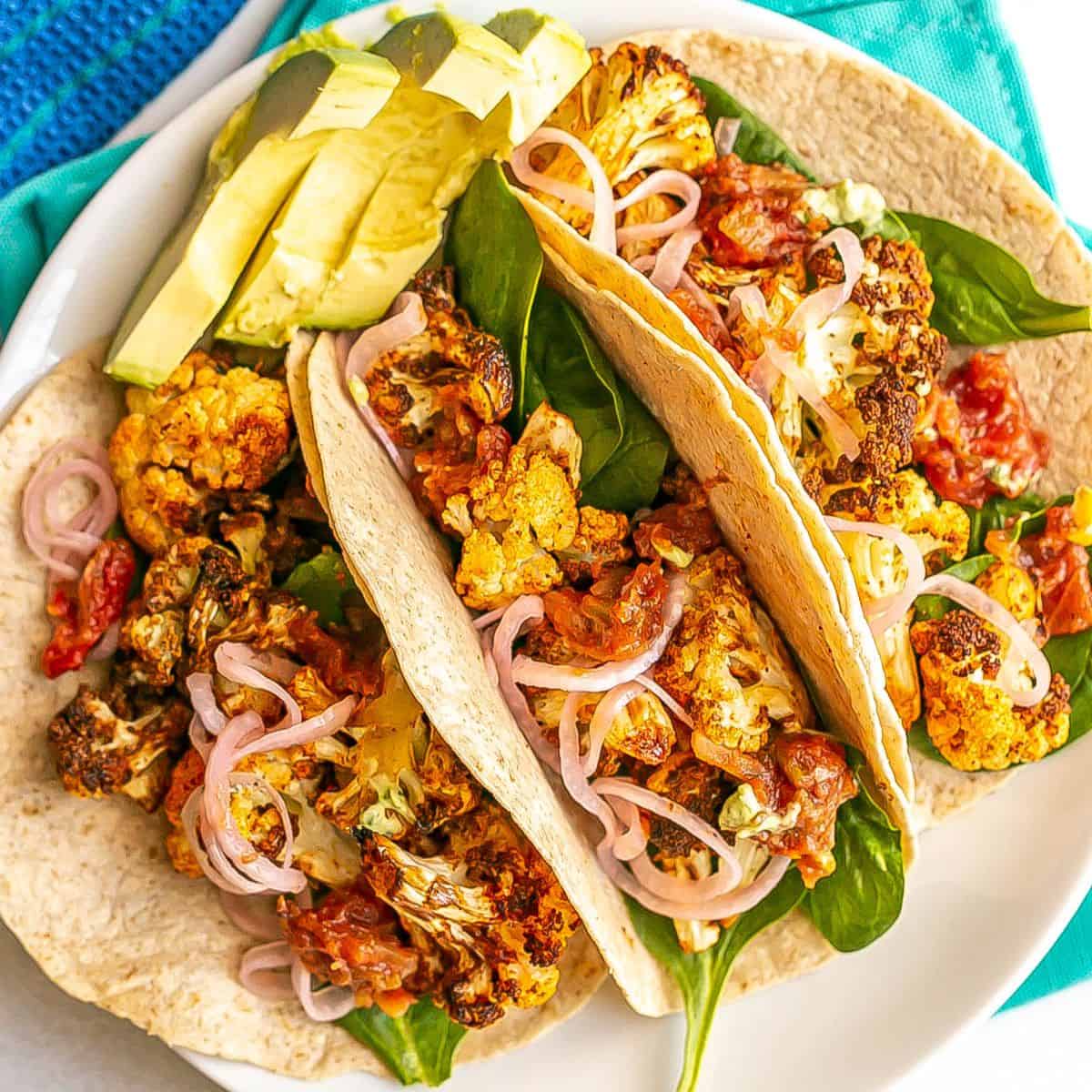 Close up of cauliflower tacos tucked together on a white plate with avocado slices to the side.