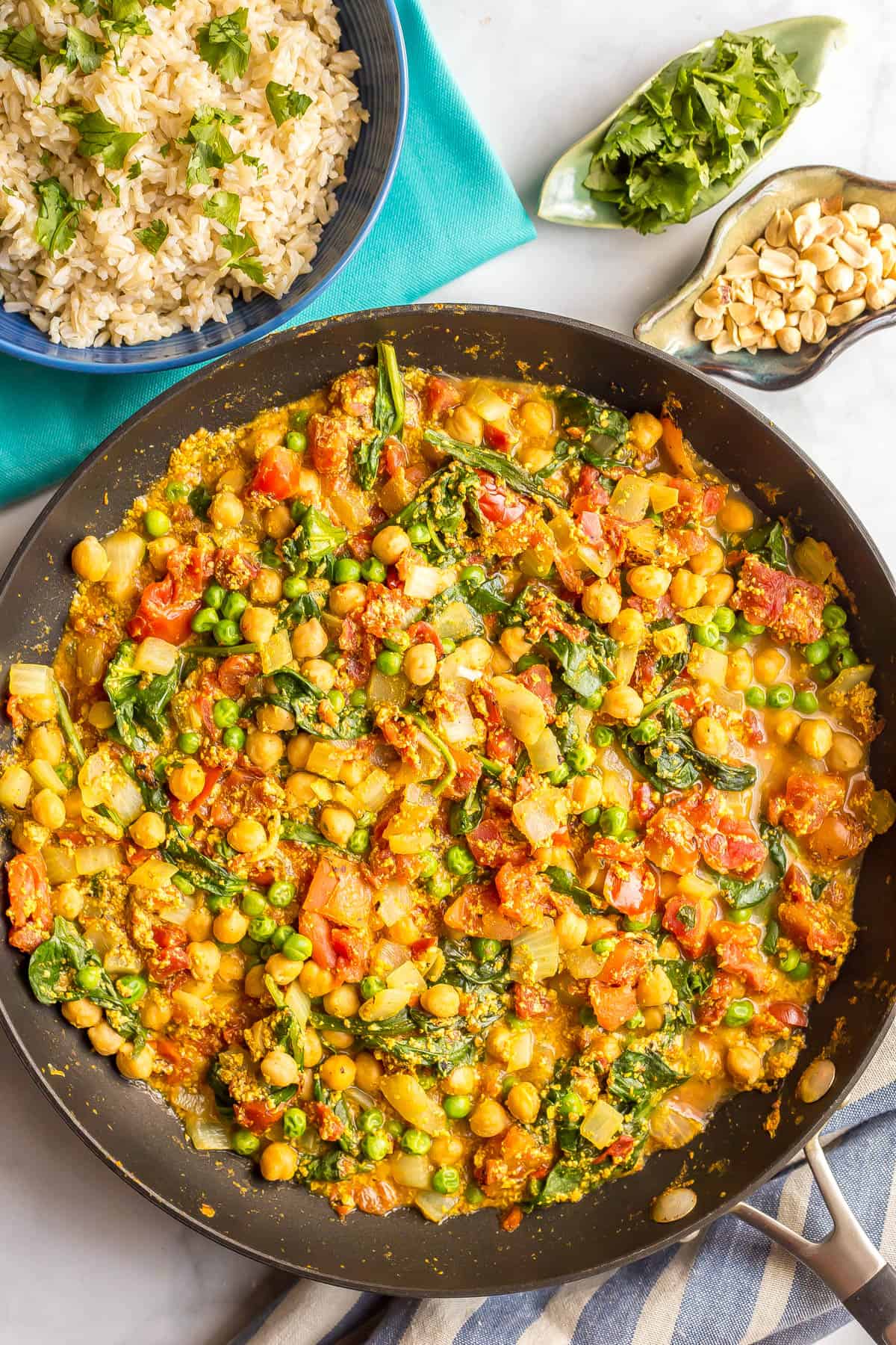 A large skillet filled with a colorful vegetarian curry of chickpeas, green peas, spinach and tomatoes in a sauce, with rice and toppings to the side.