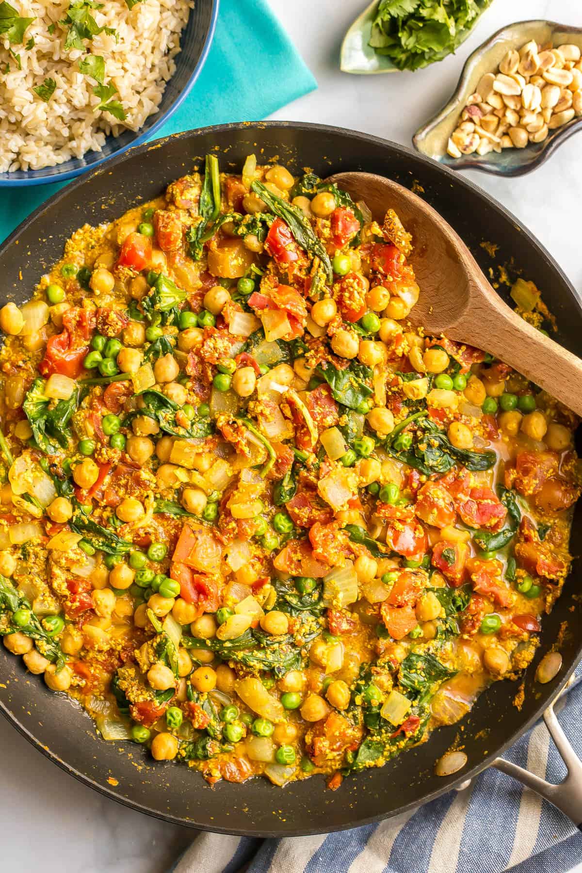 A wooden spoon resting in a skillet of a quick vegetarian curry with chickpeas, tomatoes, spinach and green peas.