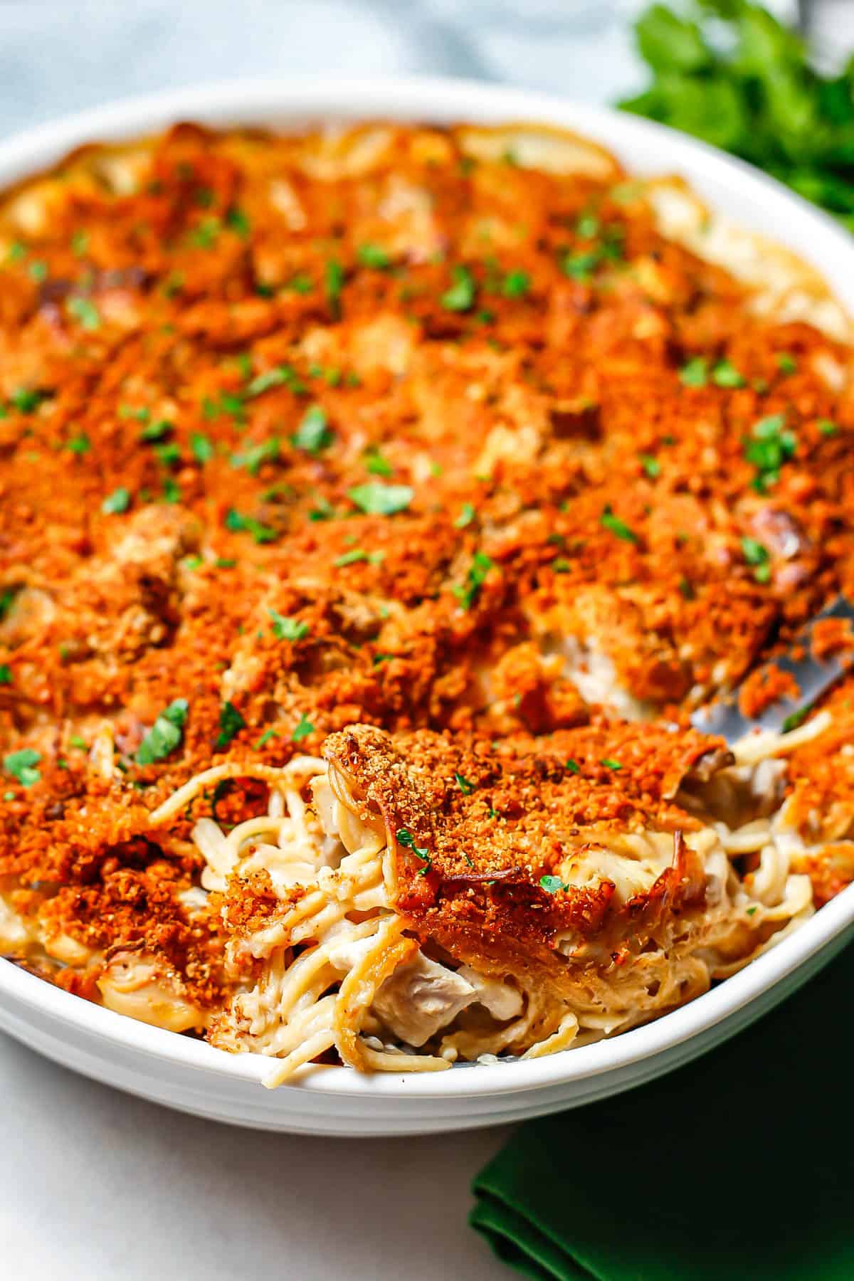 A scoop of a healthy chicken tetrazzini with a golden brown breadcrumb topping being taken from a white round casserole dish.