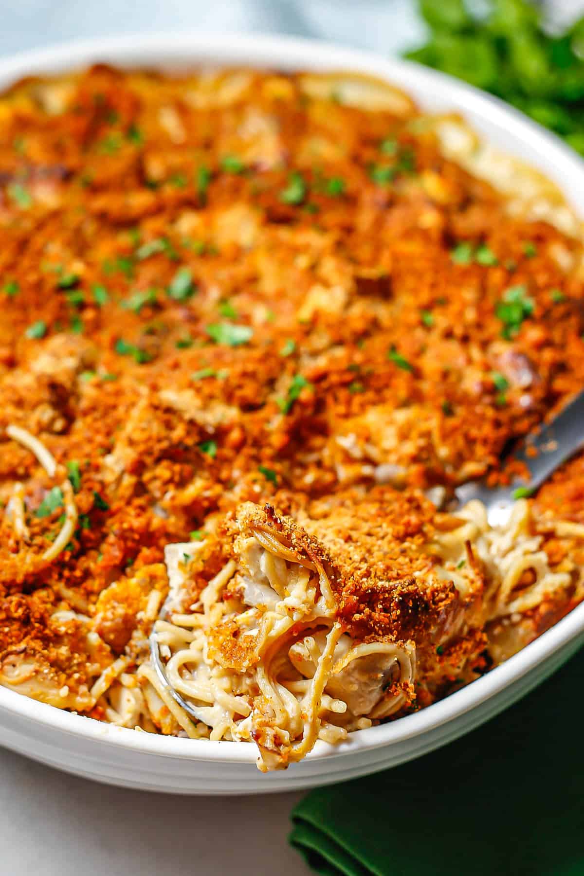 A silver serving spoon with a scoop of chicken tetrazzini with a crunchy breadcrumb topping in a large white casserole dish.