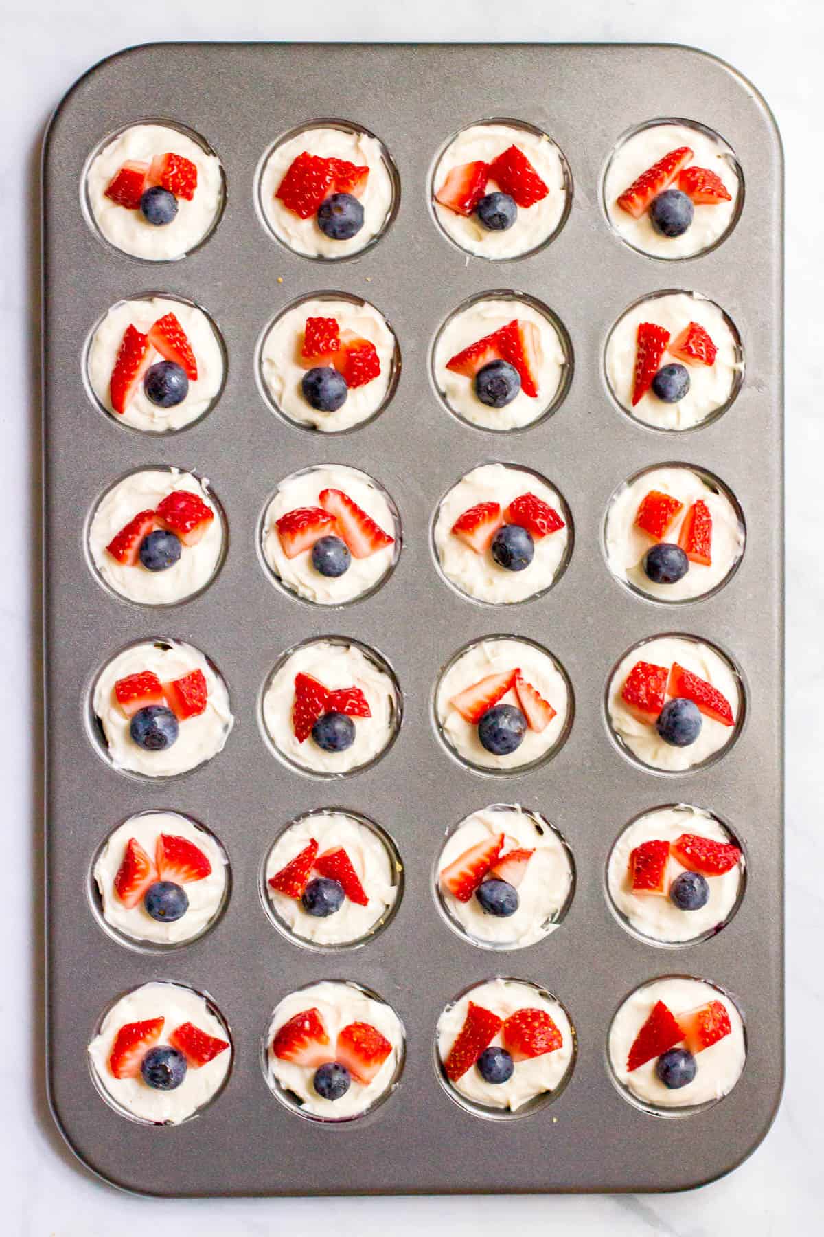 A mini muffin tin with mini cheesecake bites with strawberries and blueberries on top.