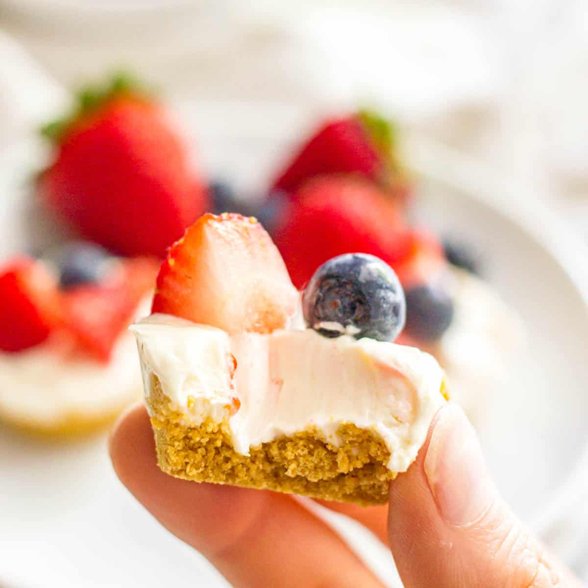 A hand holding a mini cheesecake with fruit on top and a bite taken out.