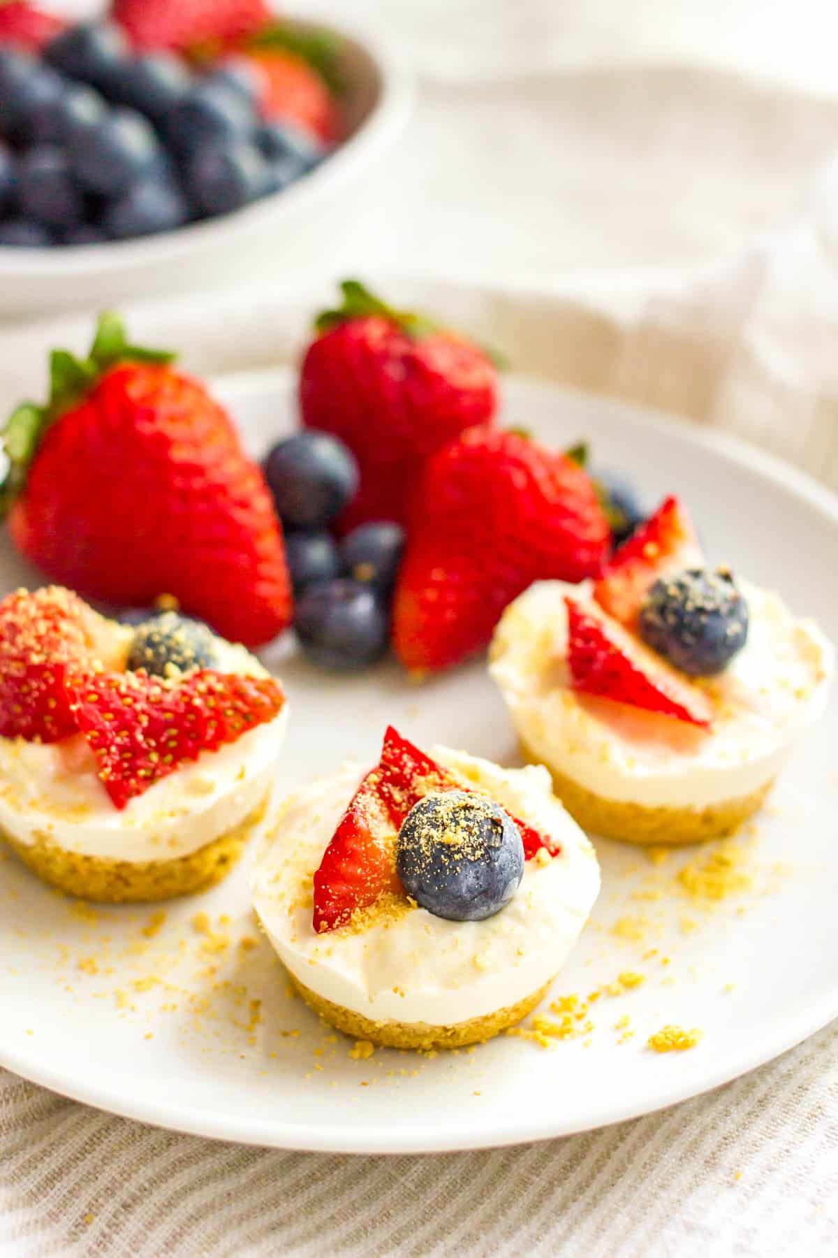 A trio of round mini cheesecake bites with a graham cracker crust and fresh fruit on a small white plate.