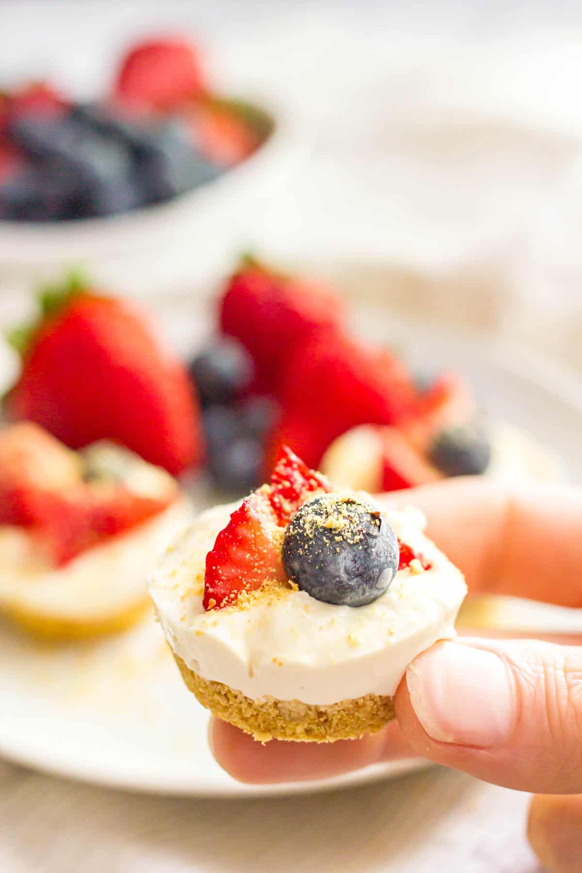 A hand holding up a mini cheesecake bite with a graham cracker crust.