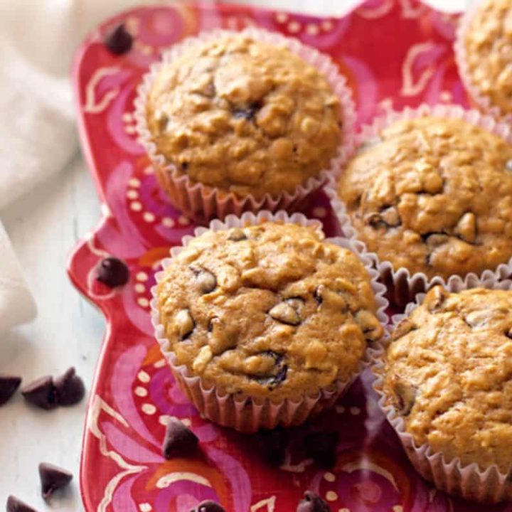 A group of oatmeal chocolate chip muffins on a red plate with chocolate chips scattered nearby.