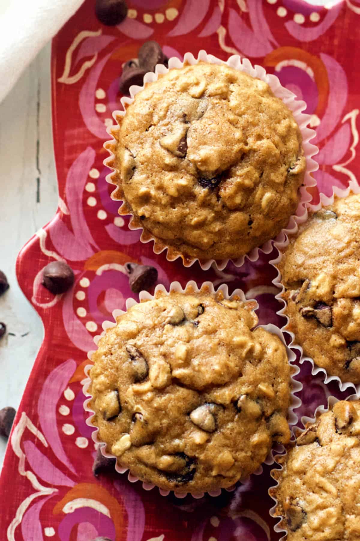 Close up of a couple of healthy oatmeal chocolate chip muffins on a red serving plate with chocolate chips scattered around.