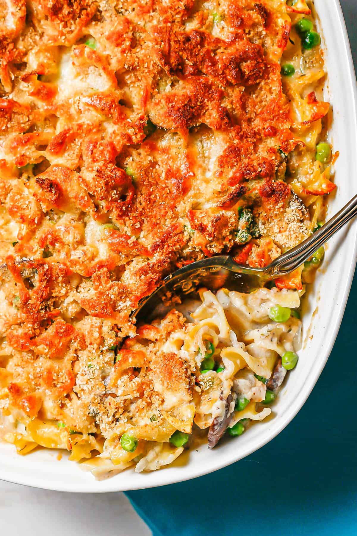 A silver serving spoon resting in a baked casserole dish of tuna noodle casserole topped with a crunchy breadcrumb crust.