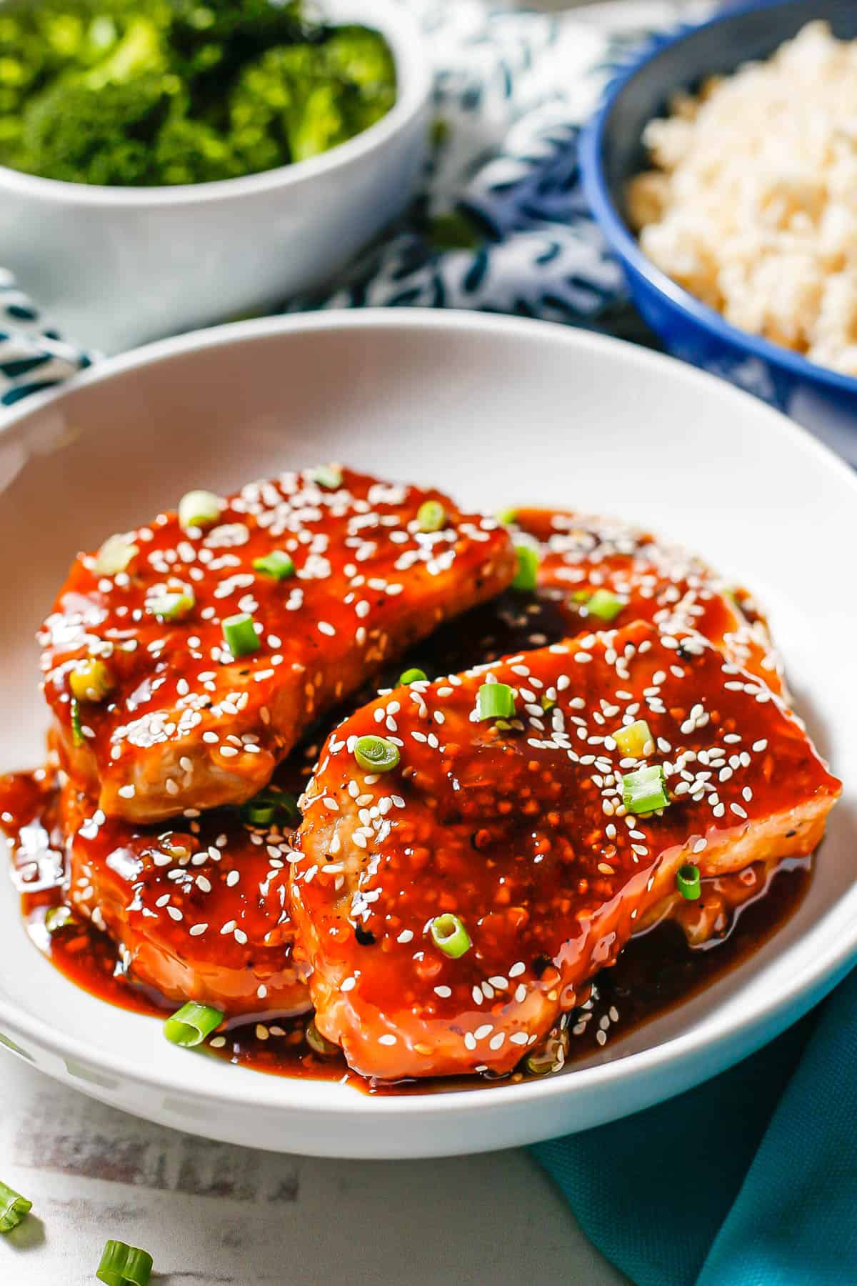 Honey garlic pork chops served in a white bowl with extra sauce with bowls of steamed broccoli and steamed rice in the background.