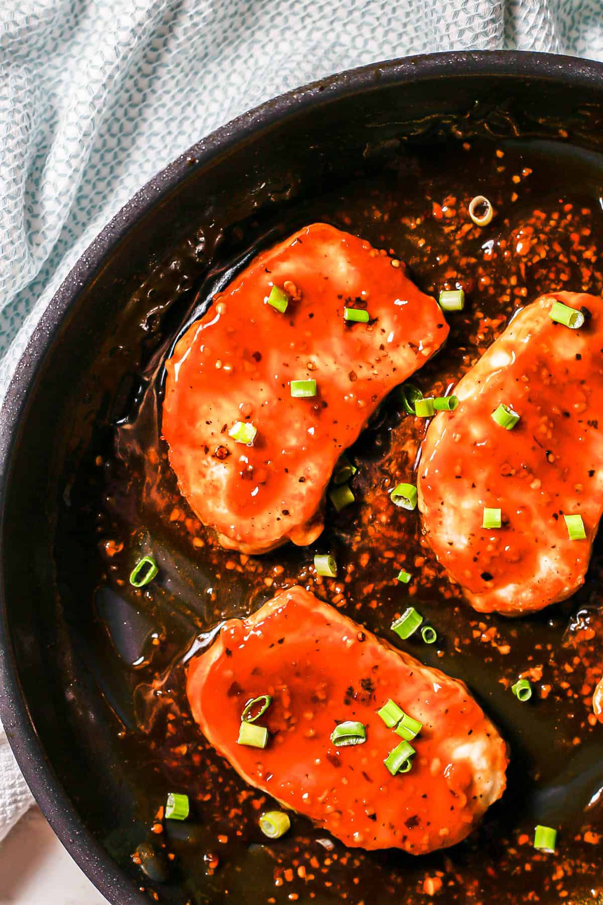 Close up of pork chops being finished in a honey garlic sauce and topped with sliced green onions in a large dark skillet.