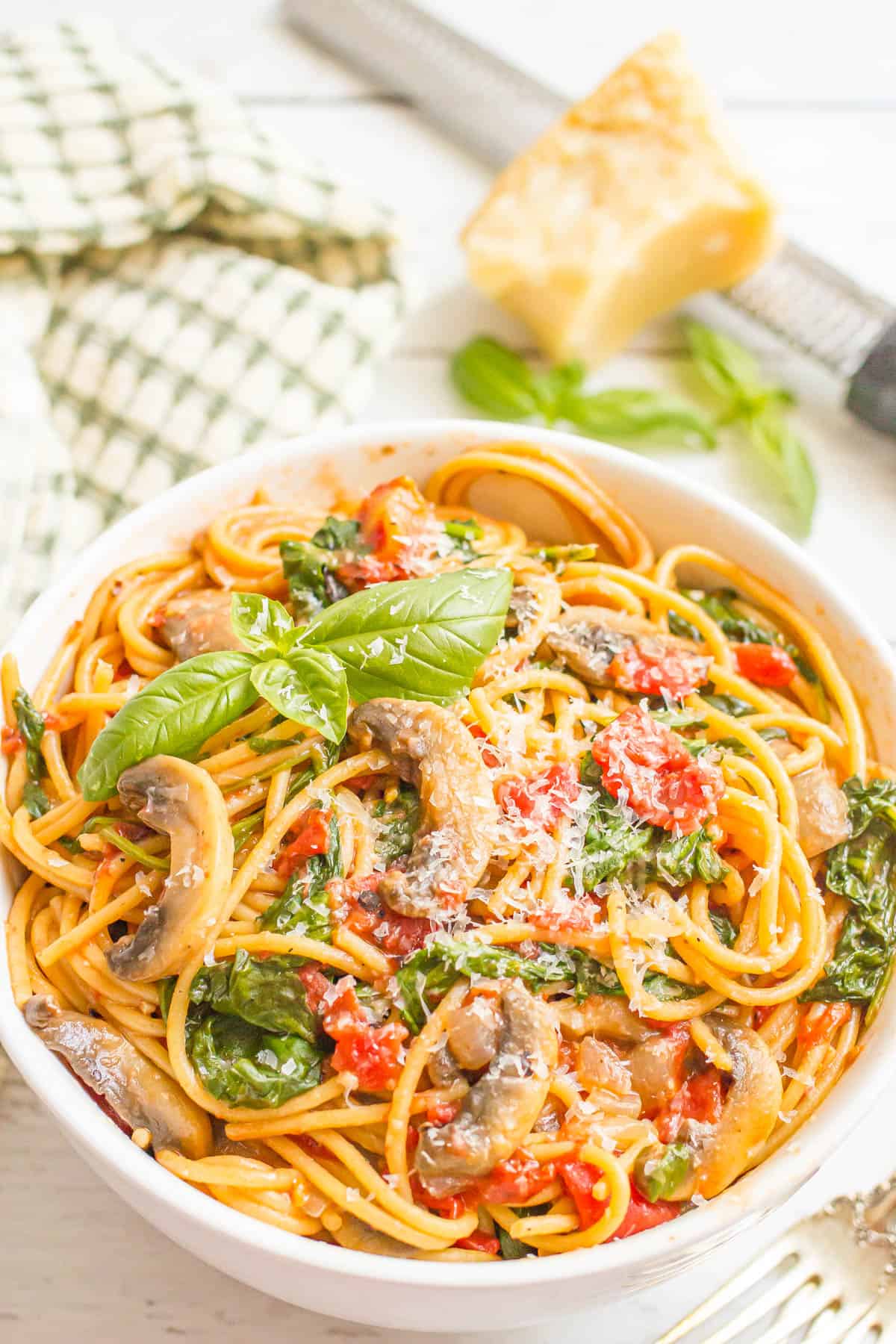 A white bowl with a vegetarian spaghetti mix including mushrooms, spinach and tomatoes with a basil sprig on top and a Parmesan rind in the background.