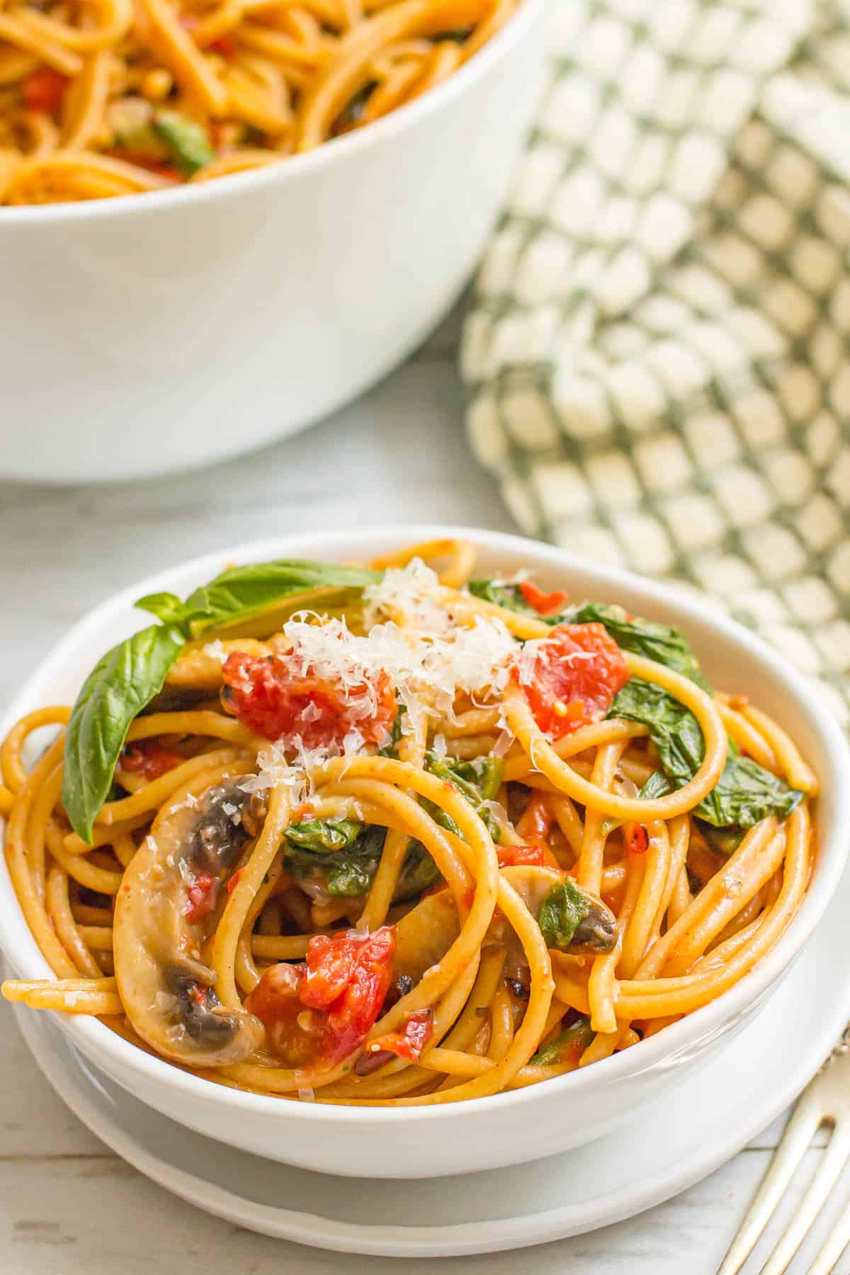 A small serving of vegetarian pasta with mushrooms, spinach and tomatoes in a white bowl set on a white plate.