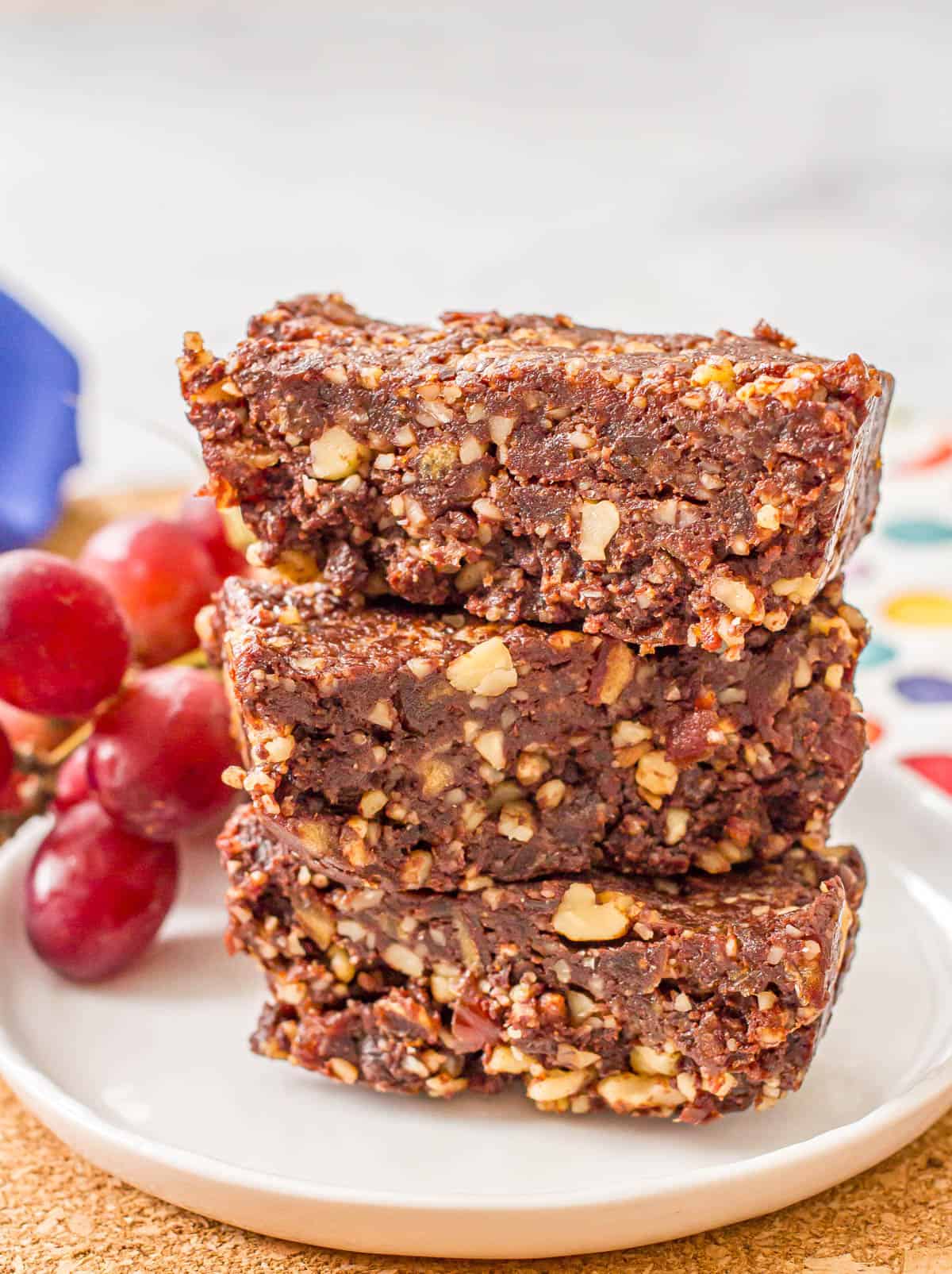 Three raw date brownies stacked on each other on a small white plate with some red grapes to the side.