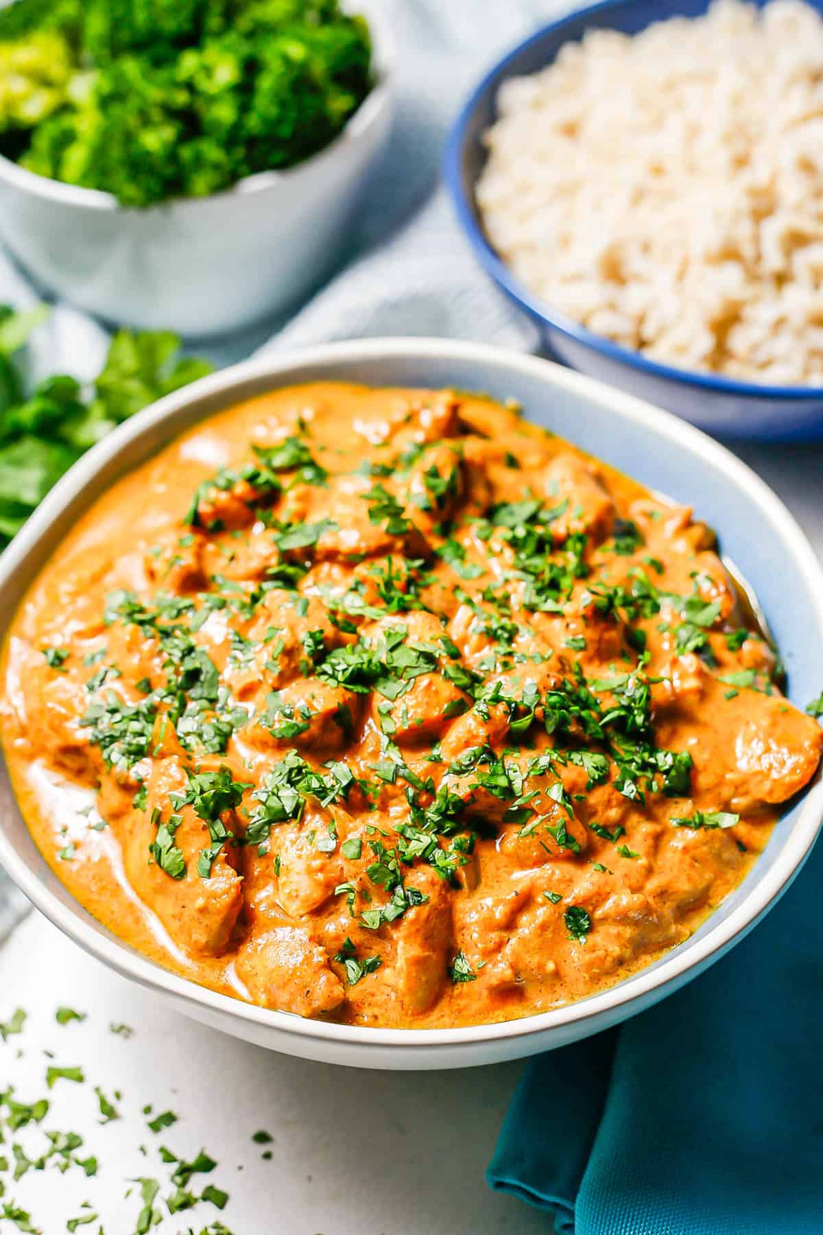 Slow cooker butter chicken served in a blue and white bowl and sprinkled with parsley, with bowls of rice and steamed broccoli in the backgorund.