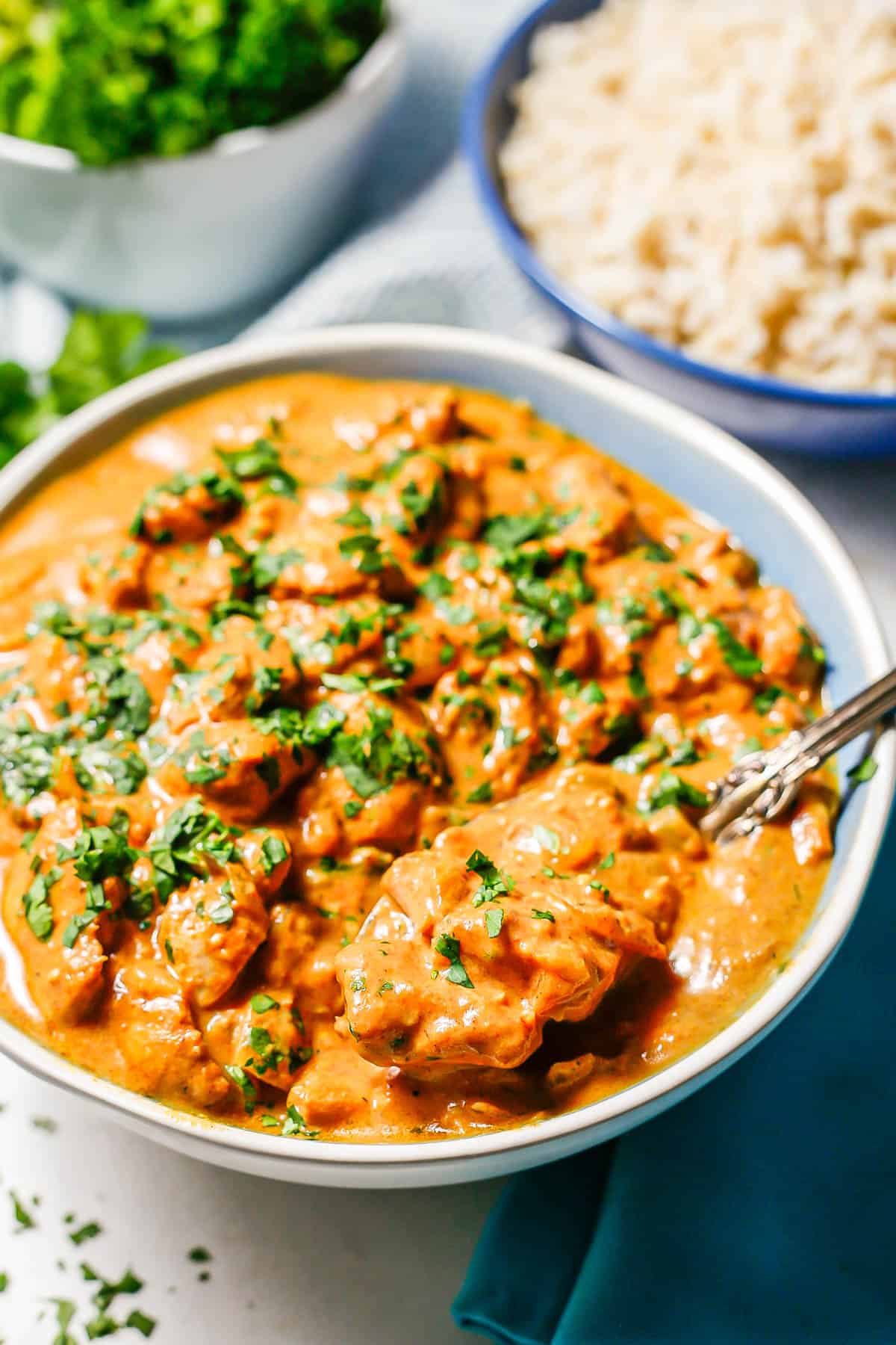 A spoon resting in a bowl of creamy butter chicken with a bowl of rice in the background.