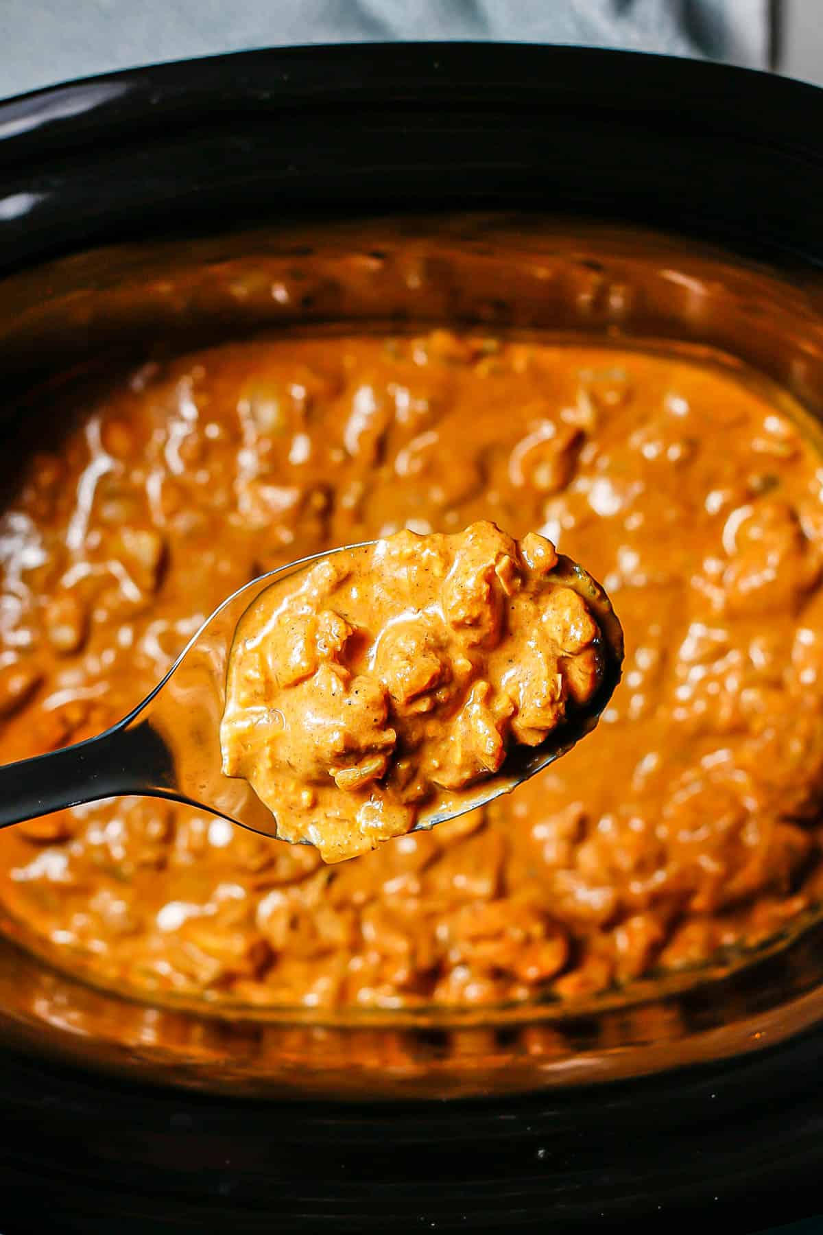 A spoon holding up a scoop of butter chicken from a slow cooker.
