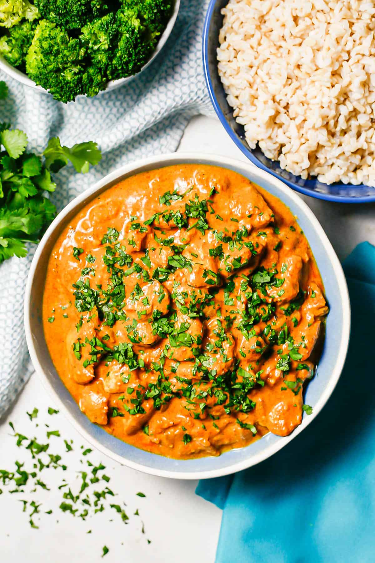 Slow cooker butter chicken served in a large blue and white bowl alongside bowls of steamed brown rice and steamed broccoli.