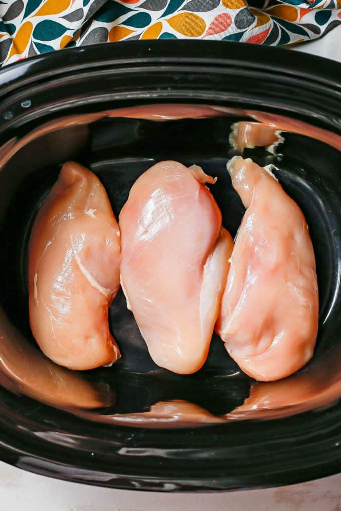 Three raw chicken breast in the bottom of a slow cooker insert.