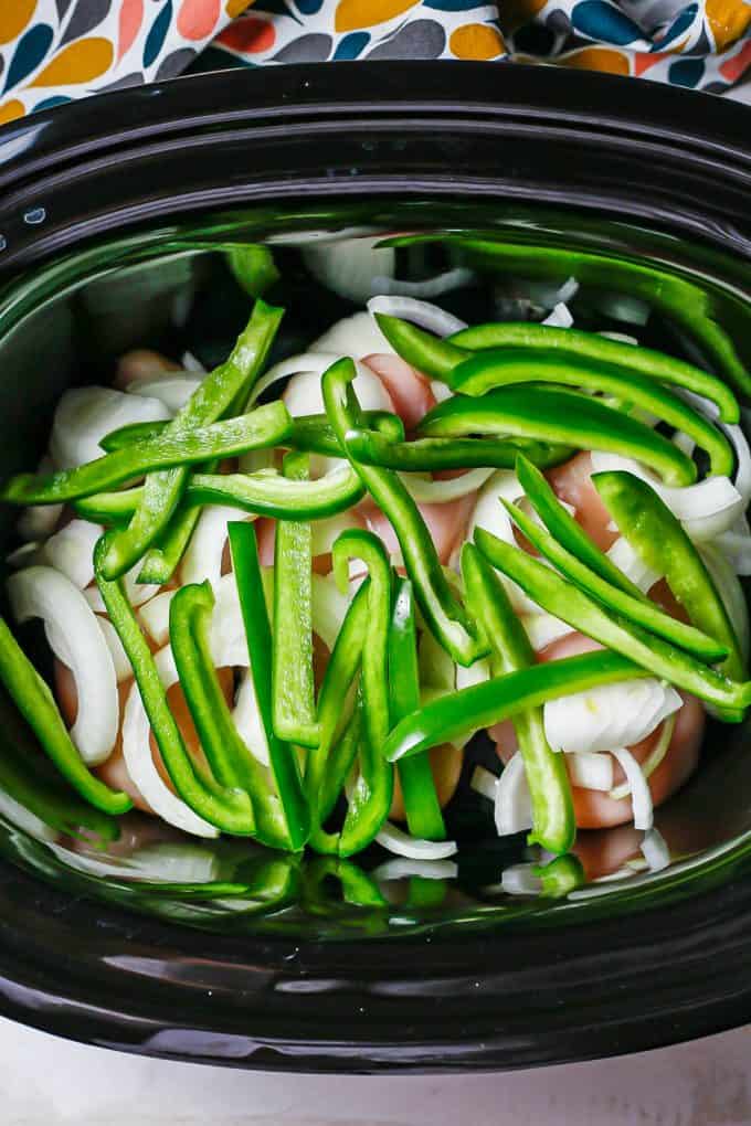 A black crock pot insert with chicken breasts topped with sliced onions and green peppers.