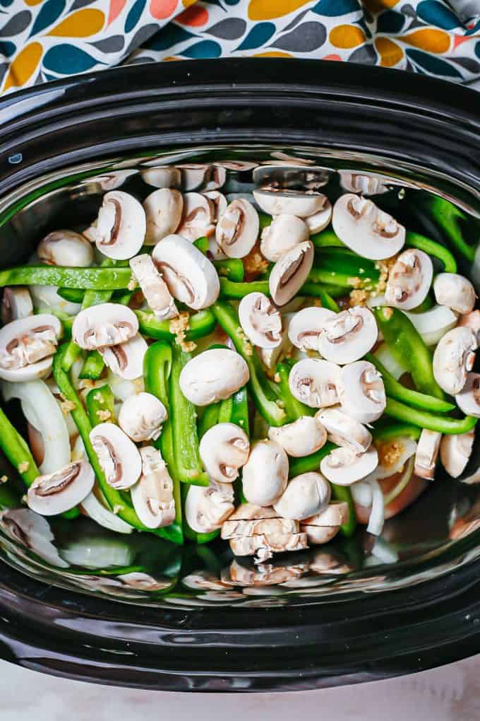 A slow cooker insert with chicken topped with onions, green peppers, garlic and mushrooms.