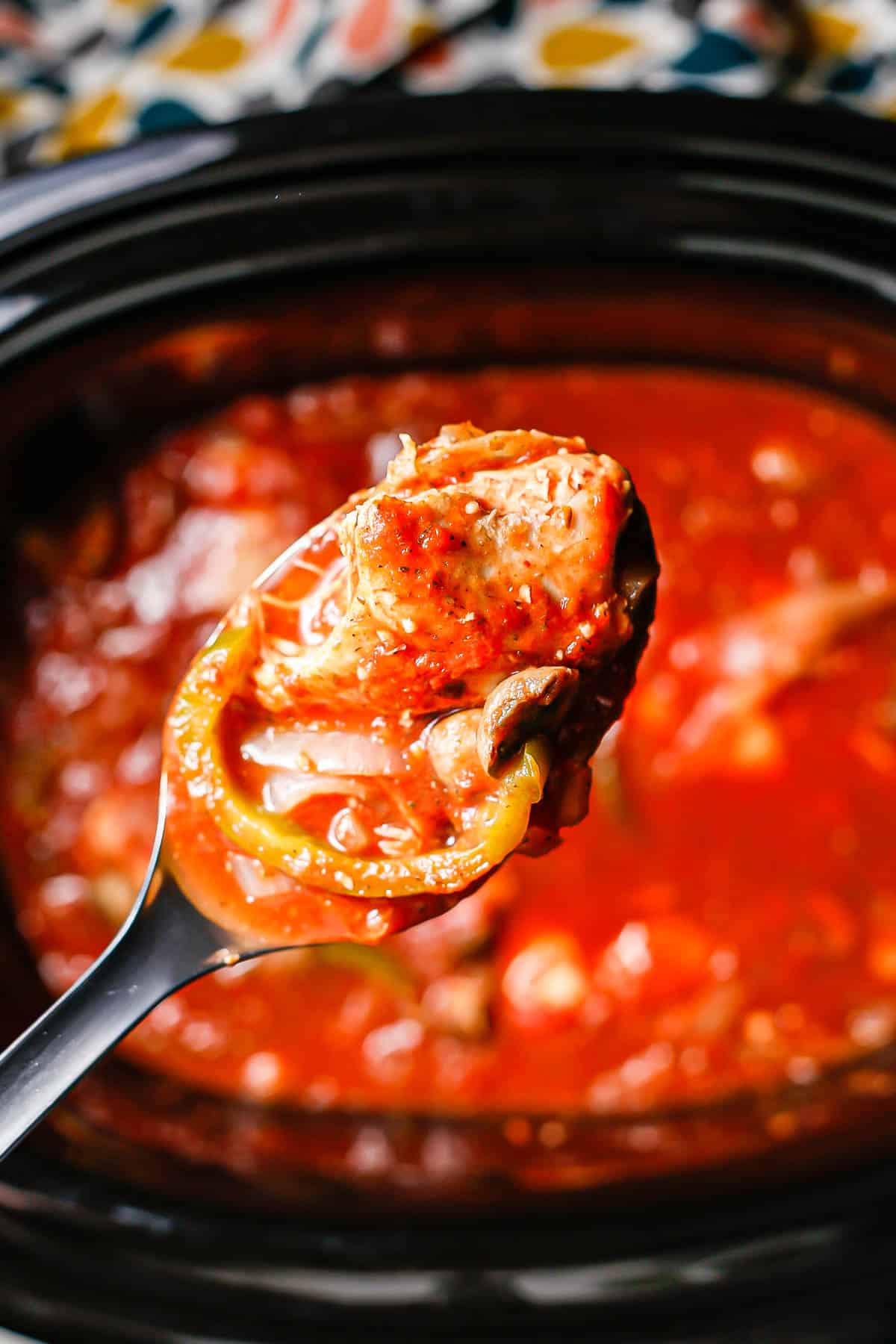 A spoon lifting up chicken and veggies in a red sauce from a crock pot.