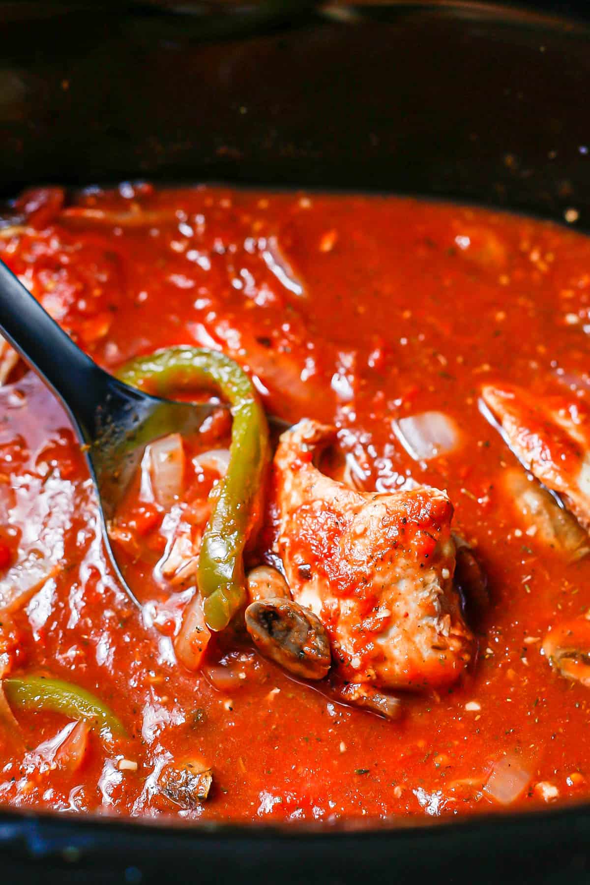 A ladle of saucy chicken resting in a crock pot with cooked chicken cacciatore.