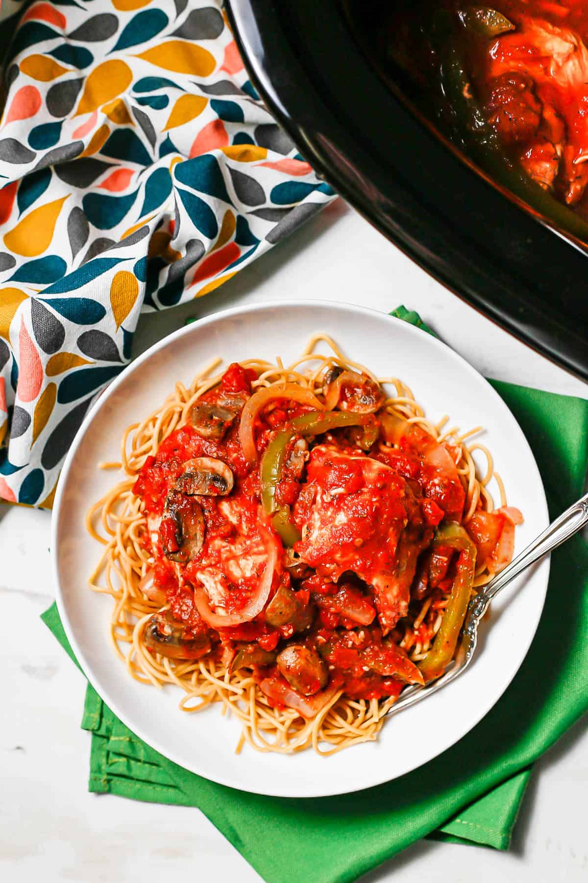 Overhead shot of a white bowl of chicken cacciatore over spaghetti beside a slow cooker of the dish.