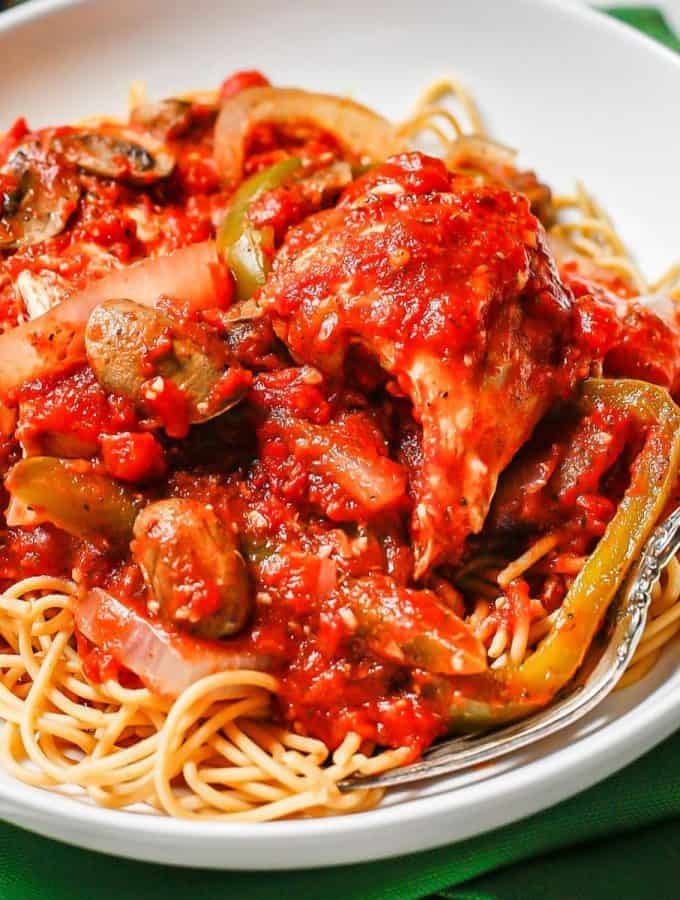 Close up of chicken cacciatore over spaghetti in a low white bowl.