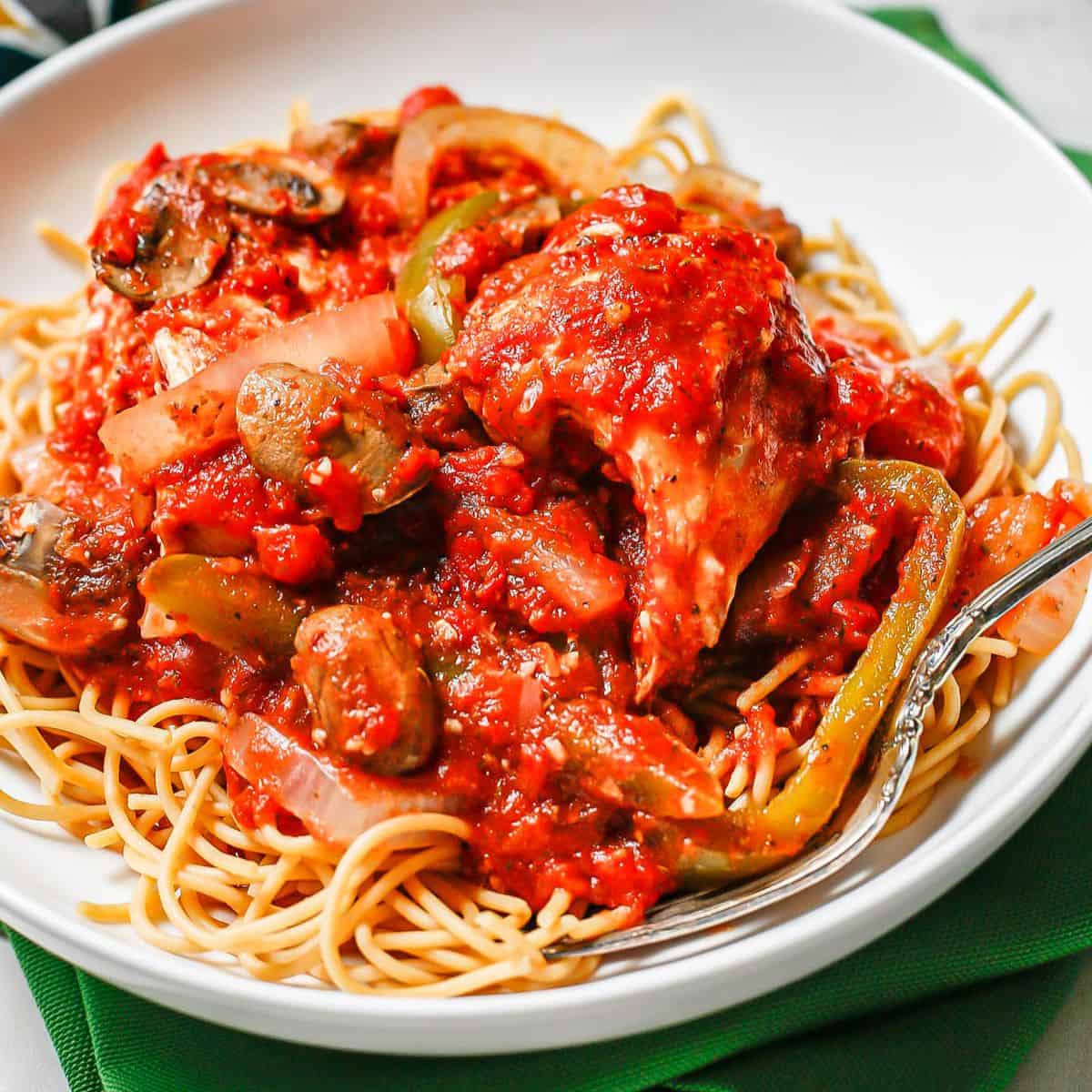 Close up of chicken cacciatore over spaghetti in a low white bowl.