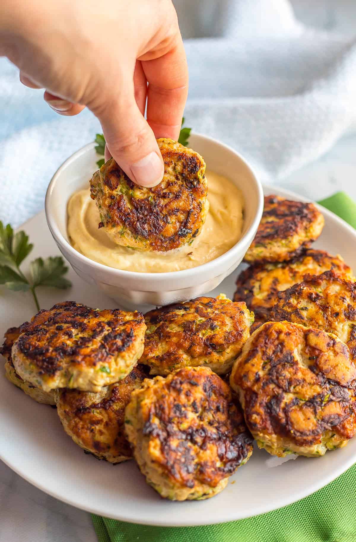 A hand dipping a spicy chicken patty into a creamy mustard mixture in a white bowl set on a white plate full of patties with green napkins to the side.