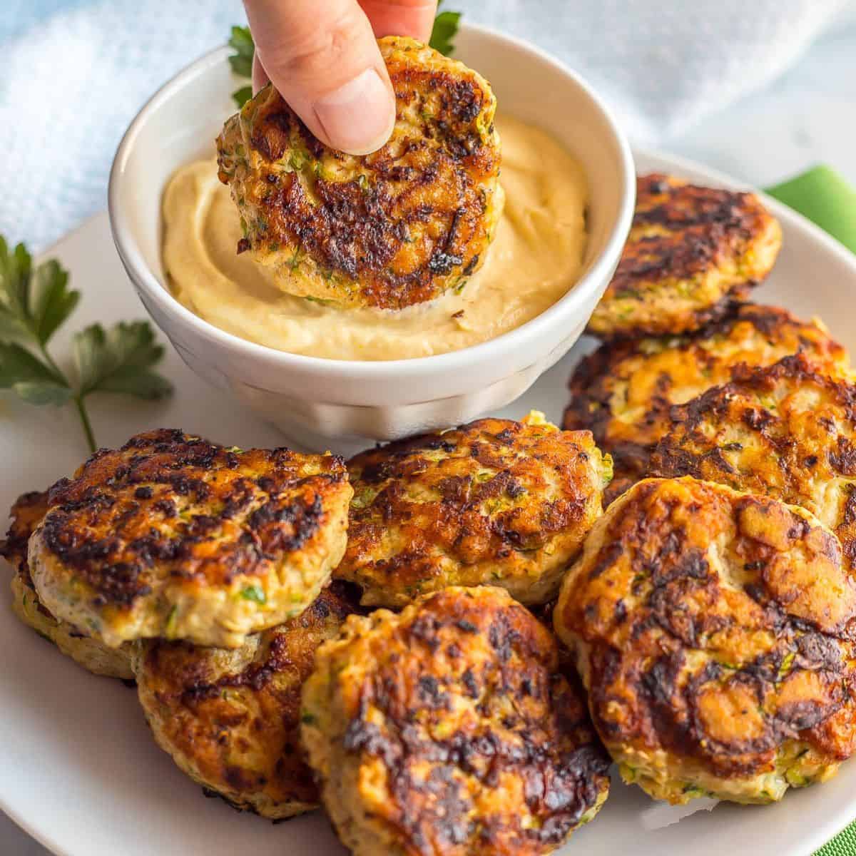 A hand dipping a spicy chicken patty into a creamy mustard mixture in a white bowl set on a white plate full of patties.