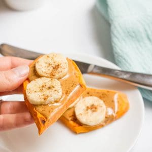 A hand holding a half of a sweet potato toast topped with peanut butter, banana slices and cinnamon.